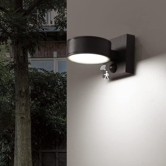 Black outdoor wall light with sensor on a gray wall, illuminated in a garden setting.