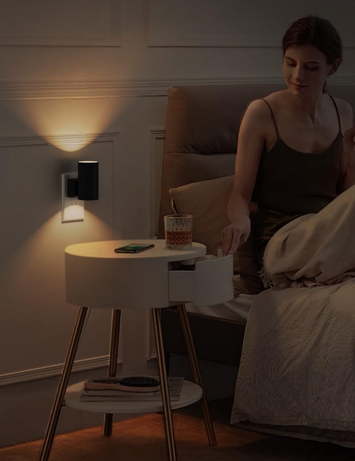 Woman sitting on a bed next to a nightstand with a lamp and glass.